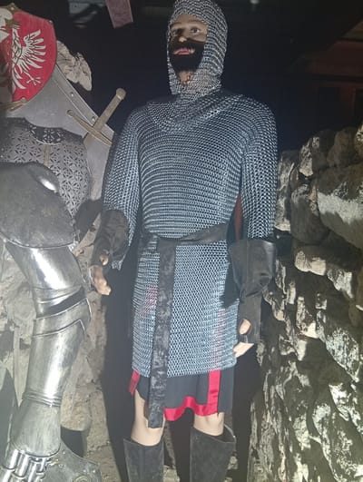 A full-body shot shows a mannequin dressed in a chainmail suit, complete with a hood, black gloves, a black belt, and a red-trimmed black skirt. The mannequin stands against a backdrop of rough stone and appears to be part of an exhibit in the city of Sobótka, Poland. To the left, a partially visible suit of silver armor and a red shield with a white eagle emblem are displayed. The lighting is dim, casting shadows and highlighting the textures of the armor and the stone.