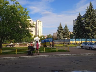 A bright, sunny day in Vyshhorod, Ukraine, is depicted in this image. Tall apartment buildings and evergreen trees dominate the skyline, with a clear blue sky and scattered clouds. In the foreground, a woman in a pink shirt pushes a stroller down a sidewalk bordering a park. She walks past benches where other people are seated and a yellow fence that separates the park from the sidewalk. Behind the fence, a monument and a series of posters featuring portraits are visible. The posters have Ukrainian flags displayed on them, and some text is visible, though it's largely illegible in the image. A silver car is parked on the street next to the park. The overall atmosphere appears calm and pleasant.