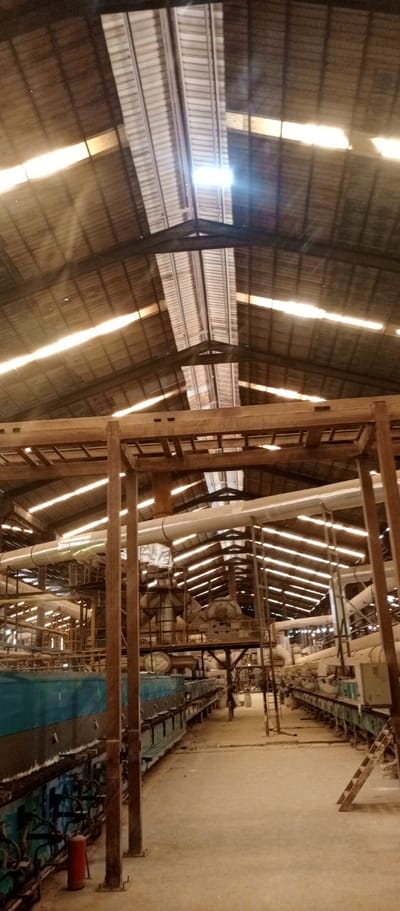 The image displays the interior of a large industrial facility or factory, likely located in Magboro, Nigeria. The perspective is looking down a wide central aisle, offering a view from the floor towards the high, vaulted roof.
The ceiling is constructed from corrugated metal sheeting, punctuated by numerous translucent skylights that allow abundant natural light to penetrate the space. These skylights create bright, almost glaring streaks of light across the roof structure, indicating it is daytime. Heavy metal beams and trusses support the roof.
Below the roof, an intricate network of industrial machinery and infrastructure is visible. Large, insulated pipes, possibly for conveying materials or air, run horizontally and vertically, supported by extensive metal frameworks and platforms. On the ground level, two long, parallel lines of industrial equipment define the central aisle. The machinery on the left consists of large, rectangular units primarily in blue-green, while those on the right are lighter in color. A red fire extinguisher is placed on the floor near the blue-green machinery on the left side. Various smaller pipes, electrical conduits, and support structures are integrated throughout the machinery. The floor is made of concrete and appears dusty.
No people are present in the image, and no specific activity or event is actively taking place, though the setup clearly indicates an operational industrial environment. There is no visible text discernible within the frame.