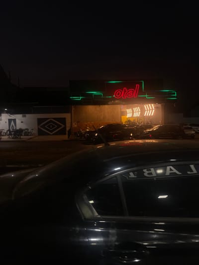 The image displays an urban street scene at night in Anápolis, Brazil. In the immediate foreground, the dark-colored roof and upper side of a vehicle are partially visible, reflecting artificial light sources and a reversed, capitalized text fragment reading "LABT" on its rear passenger window.

The midground reveals multiple vehicles, including several cars and motorcycles, parked or stopped on a paved surface, partially obscured by the darkness.

In the background, a commercial building is prominently illuminated. It features a red neon sign displaying the text "otal" on its upper facade, flanked by horizontal green neon strip lighting. Below this sign, the building's entrance is brightly lit by multiple vertical, amber-toned strip lights, which reveal an interior space containing exercise equipment. To the left of this main building, a white wall is visible, adorned with a black stylized flame graphic and a larger black diamond-shaped motif with a central circular element. Several dark outdoor tables and chairs are arranged in front of this wall. The sky above is dark, indicating nighttime conditions. No discernible individuals are present within the frame.