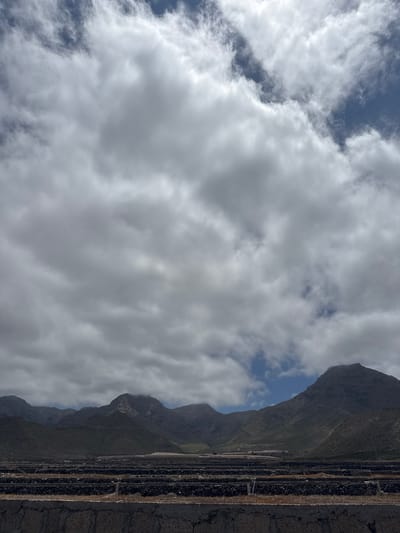 The image shows a landscape view in Adeje, Spain, with agricultural terraces in the foreground and mountains in the background. The terraces are constructed with dark stones and appear to be arranged in rows, possibly for growing crops or for water management. The mountains are rugged and arid, with some vegetation on their lower slopes. The sky is partly cloudy, with large white clouds dominating the upper portion of the frame. The overall scene depicts a rural or semi-rural area, likely a farming region within the mountainous terrain of Adeje.