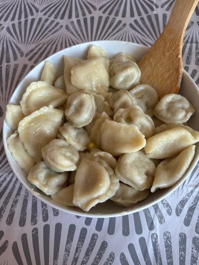 A white bowl filled with dumplings is placed on a table with a grey and white patterned tablecloth. A wooden spatula rests on the right side of the bowl, partially submerged in the dumplings. The dumplings appear to be cooked and glistening, suggesting they are either buttered or in a sauce. The lighting is bright, and the image is a close-up shot, focusing entirely on the food and the bowl. The scene is indoors. There is no information or visual evidence to determine the location in the city of Cork, Ireland.