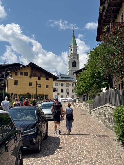 This is a vertical, eye-level, full shot of a street scene in Cortina d'Ampezzo, Italy, bathed in bright sunlight. The image is oriented sideways, with the sky appearing on the left. A prominent church steeple with a green roof and a clock face dominates the upper left portion of the frame, rising above light-colored buildings with numerous windows and a few dark shutters.
The street itself is paved with cobblestones, and several cars are parked along its edge, including a dark sedan in the foreground. A few people are visible, some walking along the street and others appearing to be conversing or engaged in activities around the cars. One person, viewed from behind, is wearing a blue shirt, dark pants, and a dark bag slung over their shoulder. Another person, also from behind, has on a backpack and dark shorts. Their postures suggest they are either walking or looking down at something.
The buildings on the right are made of a yellowish stucco with white trim around the windows. Some of the buildings have wooden architectural elements. Lush green trees with dense foliage are interspersed throughout the scene, providing shade and natural beauty. The sky is a vibrant blue with scattered white, fluffy clouds, indicating fair weather. The presence of the church steeple and the architectural style of the buildings are strong cues to the European location, specifically Cortina d'Ampezzo. The bright sunlight and clear sky suggest it is daytime, likely late morning or early afternoon. The atmosphere appears relaxed and leisurely, with people enjoying the pleasant weather and surroundings. There is no visible text in the image.