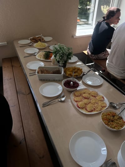 A long table is laden with food and place settings in what appears to be a communal dining area or canteen. The table is positioned near a window that offers a view of greenery and trees, suggesting an outdoor or semi-outdoor setting, possibly in a rural or suburban area. The table itself is made of light-colored wood and is covered with various dishes of food, including what looks like mashed potatoes, salad, sliced tomatoes with cheese, bread, and a rice dish. Several white plates, cutlery, and bowls are arranged along the table, indicating a meal is about to be served or is in progress.
Two individuals are partially visible in the image. One is a woman with dark hair tied back, wearing a black t-shirt and patterned skirt, seated on a wooden bench. She is looking towards the window, her profile visible. Another person, wearing a white t-shirt, is seated to the right of the woman, with only their shoulder and arm visible. The background shows a textured wall and a window with sunlight streaming in. The overall atmosphere suggests a casual mealtime setting. The location is identified as Завалочыцкі сельскі Савет, Belarus.