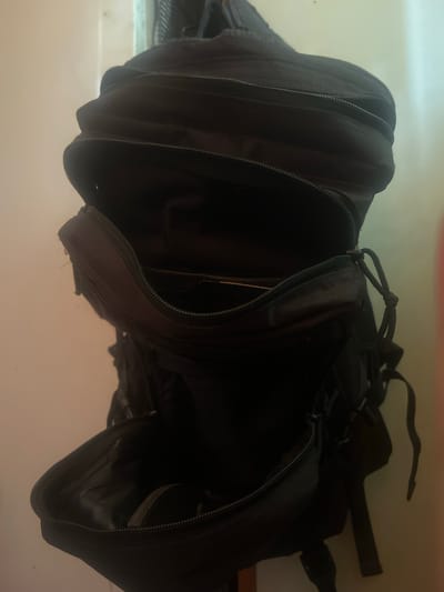 A black backpack is shown hanging against a wall, possibly in a room in Charentsavan, Armenia. The backpack is partially open, revealing its interior compartments. The lighting is dim, casting shadows and highlighting the textures of the fabric and zippers. There are no people visible in the image.