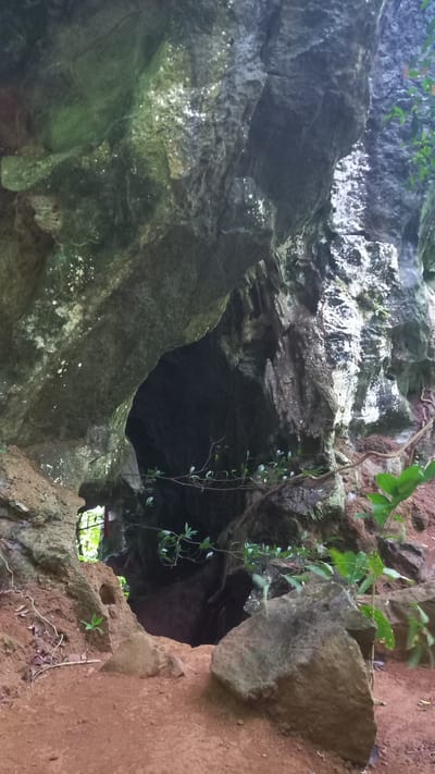 The image shows a partial view into a cave or rock shelter located in Ban Pak Ra, Thailand. The entrance is framed by uneven, textured rock formations. The left side of the entrance is predominantly gray-green with moss and lichen growth. The right side of the entrance is composed of reddish-brown earth. A few green vines with small leaves dangle from the upper right. The interior of the cave is dark, with a small opening in the back, revealing green foliage and daylight.
