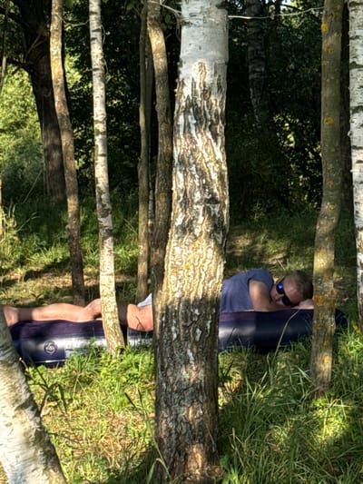 In the city of Kuzemkino, Russia, a man is seen lying on an inflatable mattress in a wooded area, resting on his side. He is wearing a blue t-shirt and dark sunglasses. The mattress is dark blue and appears to be partially deflated. The surrounding environment is characterized by a dense growth of trees with visible trunks and foliage, and lush green grass. Sunlight filters through the trees, creating patches of light and shadow on the ground. The image captures a moment of relaxation in a natural outdoor setting.