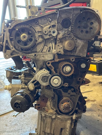 An internal combustion engine is depicted in a detailed, close-up view, its metallic components covered in dirt and grime, suggesting a state of disrepair or disassembly. The engine block, composed of various gears, pulleys, and metal casings, is the central subject. The lighting in the image is uneven, with some areas brightly illuminated and others cast in shadow, contributing to the gritty aesthetic. The background is blurred, but hints of a workshop environment can be discerned, with tools and other equipment vaguely visible. The engine itself appears to be a complex piece of machinery, with intricate interconnections between its various parts. The overall impression is one of a utilitarian and somewhat neglected industrial setting. The image was taken in Dub, Montenegro.