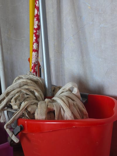 The image depicts an indoor scene featuring cleaning implements. A red plastic bucket, with a black handle mechanism on its upper left rim, occupies the lower central and right portion of the frame. Inside the bucket, a light-colored, used fabric mop head, consisting of numerous tangled strips, rests. Several poles or handles lean against a textured wall in the background. From left to right, these include a partial white pole, a bright yellow pole, a red pole adorned with a white floral pattern, and a silver metallic pole. Orange and black plastic components are visible near the base of the patterned red pole, potentially part of a mop wringer. The background wall is light grey/beige, exhibiting multiple hairline cracks, an uneven surface, and subtle blue and yellow discolorations. Vertical shadow patterns are cast on the wall. No individuals are present, and no actions are occurring. The visual information within the image does not provide any verifiable details to specify the location as the city of Kyustendil, Bulgaria.