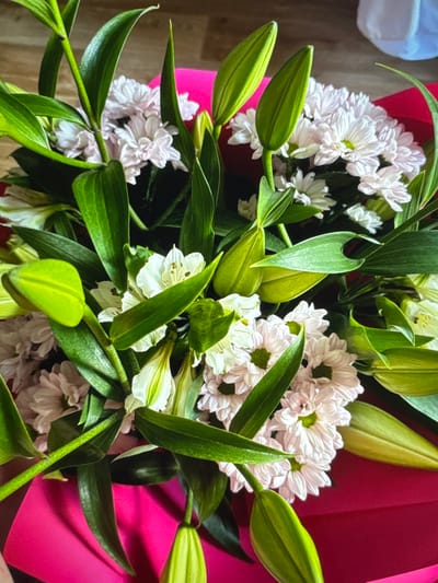A close-up, overhead shot shows a bouquet of white and pink flowers. The bouquet is arranged with vibrant green leaves and buds. The flowers are densely packed and appear to be lilies and chrysanthemums. The bouquet is presented in a bright pink wrapping. The background is blurred, suggesting the image was taken indoors with a shallow depth of field. The lighting is natural, highlighting the textures and colors of the flowers. The scene is static, with no discernable action or interaction taking place. The location is identified as Novinki, Russia.