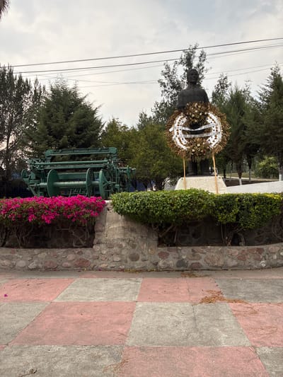 The image shows an outdoor scene in Tepeji de Ocampo, Mexico. A bronze statue stands as a central element, adorned with a large wreath. A stone monument with an inscription is positioned behind it, flanked by hedges. In the foreground, a green metal structure resembling a weaving machine sits adjacent to a stone retaining wall, partially covered in pink bougainvillea flowers. The ground is paved with a checkered pattern of pink and grey tiles. Power lines cut across the upper portion of the frame against a cloudy sky. Trees surround the scene, adding to the park-like atmosphere.