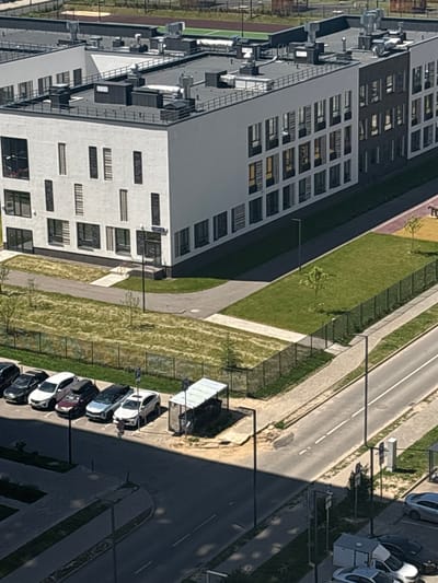 The image shows an aerial view of a modern, multi-story building with a dark and white facade, likely a school or office building, located in Misaylovo, Russia. The building is situated next to a paved road and a parking area containing several cars, including white SUVs and sedans. Adjacent to the building is a green lawn bordered by a black metal fence. Further away, a sports field with a running track is visible. The environment appears to be a well-maintained urban or suburban area. There are no people visible in the image.