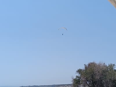 A paraglider soars through a clear, pale blue sky on a bright day. The paraglider, a small figure with a blue parachute, is positioned in the upper-middle section of the frame, creating a sense of vastness and freedom. Below the paraglider, a hint of the landscape is visible, with a line of trees and hilly terrain suggesting an outdoor, likely coastal or rural, setting. The upper right corner of the image features a portion of an architectural element, possibly a building's overhang or a white structure, casting a subtle shadow and adding a human-made contrast to the natural scenery. The overall mood is serene and adventurous, capturing a moment of aerial recreation against a backdrop of nature and a touch of human presence. The time of day appears to be daytime, given the clear sky and bright lighting. No text is visible in the image. The scene is consistent with the landscape of Episkopi Cantonment in the Sovereign Base Areas of Akrotiri and Dhekelia, a location known for its open spaces and potential for recreational activities.