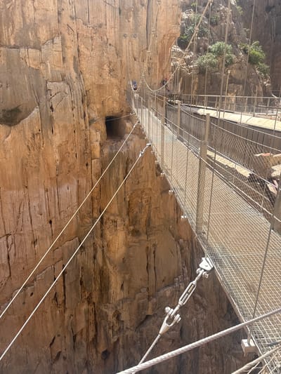 The image depicts a dramatic suspension bridge spanning a deep gorge with sheer rock faces. The bridge is constructed with a metal grid walkway and sturdy cables, showcasing impressive engineering against the natural ruggedness of the environment. The rock walls are layered and textured, indicating a geological history of erosion and time, with small patches of vegetation clinging to crevices. A person in a red helmet is visible on the bridge, adding a sense of scale and human presence. The light suggests it is daytime, likely with clear weather, casting shadows that highlight the contours of the rock. The scene evokes a sense of adventure and awe, typical of dramatic natural landscapes. The location is identified as Antequera, Spain, known for its dramatic gorges and pathways. There is no visible text in the image.