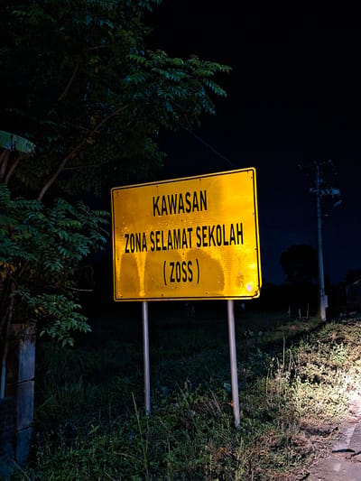 A photograph taken at night in Kutaglumpang, Indonesia, shows a yellow road sign that reads "KAWASAN ZONA SELAMAT SEKOLAH (ZOSS)". The sign is lit by an unseen light source, possibly headlights, and is positioned near vegetation. A utility pole is visible in the background. The setting suggests the sign is located near a road or path leading to or from a school.