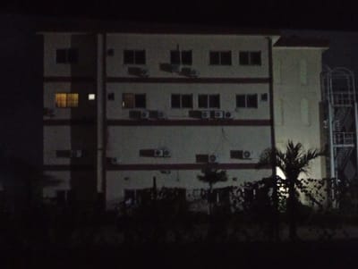A nighttime photograph shows a building in Ijanikin, Nigeria. The building appears to be a multi-story residential or commercial structure, with several windows and air conditioning units visible on the exterior walls. A fire escape is present on the right side of the building. In the foreground, there are trees and bushes obscuring the lower portion of the building. The lighting is dim, making it difficult to discern specific details of the building's facade or the surrounding environment.