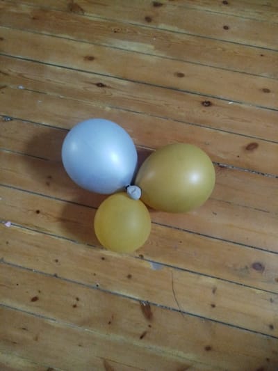Three balloons, one light blue and two yellow, are clustered together on a wooden floor. The floorboards are oriented diagonally from top left to bottom right. The balloons cast faint shadows on the wooden surface. The image is taken from a slightly elevated perspective, looking down on the balloons. There are no people or other objects visible in the scene. There is no indication of location in the city of Sabunchu, Azerbaijan.
