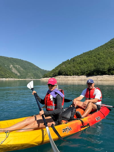 Two people are kayaking on a clear blue lake, with a lush green mountainside rising steeply from the water in the background. The sun is shining brightly, indicating it is daytime. The water is calm and reflects the sky and surrounding landscape. One person, wearing a red cap and a purple scarf over a life vest, is in the foreground, holding a paddle. The other person, wearing a blue baseball cap and a white t-shirt with a life vest, is in the background. Both kayaks are tandem, with the person in the foreground in the rear seat and the person in the background in the front seat. The kayaks are orange and yellow. The person in the background has "RTM" visible on their kayak. The person in the foreground has "RTM" visible on their kayak and a visible brand name of "OCEAN" on the orange kayak. The person in the background has the brand name "DUG" visible on their kayak. The scene suggests a recreational activity in a natural environment.