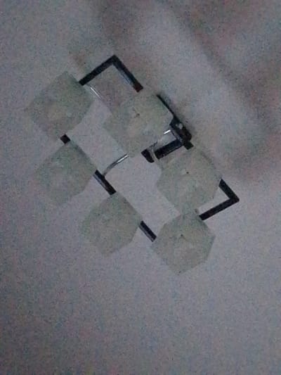 The image shows a ceiling light fixture with a modern design. It has a dark-colored frame, possibly metal, arranged in a geometric pattern. Attached to the frame are several cube-shaped lampshades, which appear to be made of frosted glass or a similar translucent material. The light fixture is mounted on a textured ceiling, which is light in color and appears to have some subtle variations in shade or pattern. The image is taken from a low angle, looking upwards at the fixture. The overall impression is one of a contemporary interior lighting element. The specific location in Yanino-1, Russia, cannot be determined from the image itself, as there are no identifying landmarks or contextual clues present.