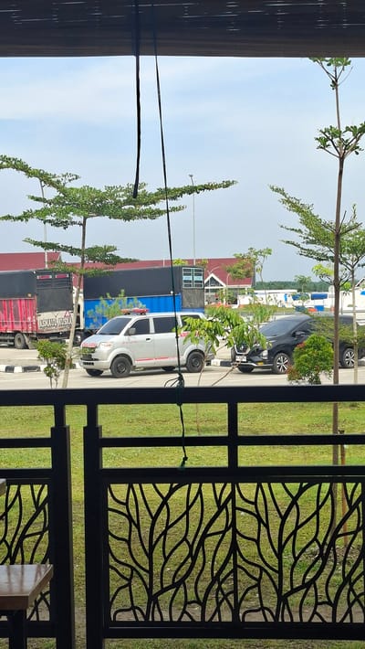 The image depicts an outdoor scene in Gohor Lama, Indonesia, focusing on vehicles and infrastructure. In the foreground, a black metal fence with a decorative leaf pattern frames a view of a grassy area. Beyond the fence, on a paved surface, a silver ambulance with red and white markings and a flashing red light on its roof is parked. To the right of the ambulance, a black minivan is also parked. In the background, two large trucks are visible; one is red and white with "ASMAUL HUSNA" written on its side, and the other is dark blue. Several trees with sparse green foliage are scattered across the scene, and in the far background, buildings with red roofs and blue and white accents suggest a commercial or rest stop area. The sky is overcast with a light blue hue. The image appears to be taken from a sheltered viewpoint, possibly a balcony or covered patio, indicated by the wooden slatted ceiling and hanging dark cords at the top of the frame.