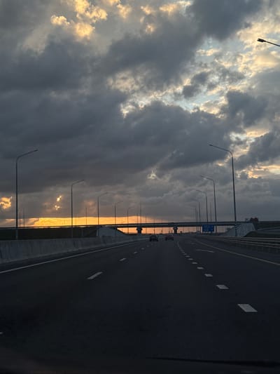 The image shows a highway in Maryanskaya, Russia, at sunset. The sky is filled with clouds, with golden light shining through. Streetlights line the side of the highway, and a few cars are visible in the distance. The road is paved and marked with white lines.
