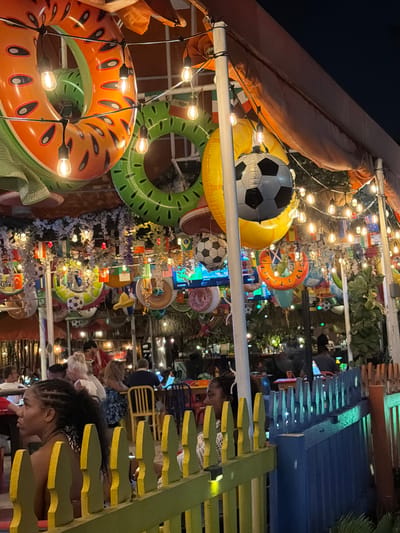 This is a nighttime, eye-level, medium shot of an outdoor dining area in Hollywood, USA, decorated with inflatable pool rings, soccer balls, and string lights, suggesting a festive or sports-themed atmosphere. The foreground features a yellow and green wooden fence. Behind the fence, patrons are seated at tables, some engaged in conversation or using electronic devices. The background includes lush greenery and more decorative elements, indicating a restaurant or bar setting. The overall lighting is dim, with string lights providing warm illumination, creating a lively yet intimate ambiance.