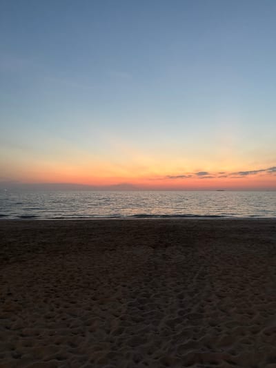 The image presents a static, panoramic view of a beach and ocean at sunset in Phra Ae, Thailand. The lower portion of the frame features dark, textured sand exhibiting numerous indentations, consistent with footprints or natural ripples. The midground is comprised of the calm, dark surface of the sea, with subtle waves visible near the immediate shoreline. On the distant left horizon, a faint light source is discernible. Towards the far right horizon, a small, dark, indistinct silhouette, consistent with a vessel or distant landmass, is present. The upper portion of the frame displays a sunset sky transitioning from a clear blue at the zenith, through a light blue and soft orange band, to a vibrant orange and reddish glow above the horizon line. Sparse, wispy clouds are present, primarily in the upper right quadrant, where a faint white point, possibly a celestial body, is also visible. No individuals, explicit actions, or direct interactions are depicted within the scene.