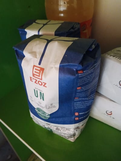 The image shows a close-up view of two bags of flour, with one bag prominently in the foreground. The bag in the foreground is blue and white, with the brand name "E'ZOZ" in red and white lettering. The text on the bag is in Uzbek and Russian, indicating the flour is "OLIV NAVLI UN," which translates to "High-grade flour." There is also an emblem of wheat stalks and a "100% Natural" seal. Another bag of flour, white with black lettering, is partially visible behind the primary bag. The bags are placed on a green shelf, and a partially visible bottle with an orange liquid is in the background. The image does not provide any specific location details within Shurchi, Uzbekistan, or any context related to people, actions, or interactions.