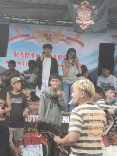 The image depicts a group of people in what appears to be an outdoor event, likely in Pendem, Indonesia, based on the Indonesian flag and Garuda Pancasila emblem in the background. There are two people on a makeshift stage holding microphones and addressing the crowd. Several individuals are gathered around the stage, some smoking cigarettes. The environment seems casual and informal, with a banner overhead displaying the words "KARANG TARUNA NUSA." Another banner, possibly related to "PAMILU COKLAT", is visible hanging above the stage on the right side. The overall scene suggests a community gathering or local performance.