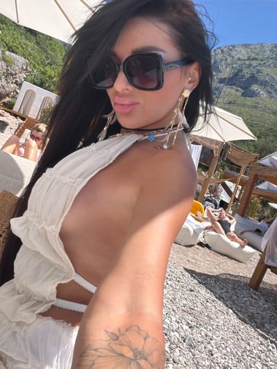 A woman, facing the camera with her head turned to the side, is taking a selfie on a pebble beach. She has long, dark hair, large black sunglasses, and is wearing a white, ruffled dress. She is adorned with a necklace and long, dangling earrings with feathers and beads. To her left, a woman with blonde hair is lying on a lounge chair, looking at her phone. In the background, there are several white umbrellas, beach structures, and people relaxing on beanbag chairs. The scene is set against a backdrop of a rocky, green mountainside and a clear blue sky. The location is Drobnići, Montenegro.
