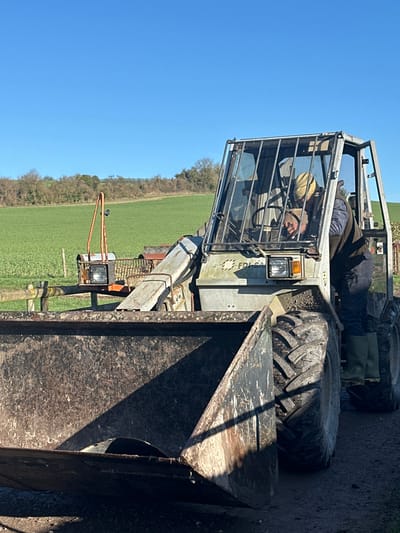The image presents an outdoor rural scene in Broad Chalke, United Kingdom, under a clear, bright blue sky. A weathered, grey-beige front-end loader, bearing the inscription "EDIL SA" on its front chassis, occupies the right and central foreground. The loader is equipped with a large, dark, and visibly dirty metal bucket positioned low to the ground. Inside the protective barred cab, a single individual is partially visible, wearing a yellow beanie, dark upper garments, and green Wellington boots. The person appears to be leaning forward, engaging with the internal controls or steering wheel. The machine is stationary on a dirt or gravel surface. In the mid-ground to the left, a portion of another piece of rusty orange farm machinery, featuring a single headlight and a tall, triangular frame, is discernible behind a rustic wooden fence post. Beyond the immediate area, vibrant green fields stretch towards a distant tree-lined ridge under strong natural lighting that casts distinct shadows.