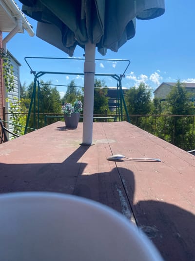 In Bol'shoe Zaborod'e, Russia, an outdoor scene unfolds under a clear blue sky with scattered white clouds. The foreground is dominated by a weathered, reddish-brown wooden table, its surface showing signs of age and use. A white umbrella pole stands vertically in the center of the table, casting a prominent shadow. To the left of the pole, a gray planter holds artificial purple and green flowers. Further to the right on the table, a single silver spoon rests, casting its own shadow. The middle ground reveals a green metal swing set. Beyond the swing set, a row of lush green trees forms a natural backdrop, with houses visible through the foliage. The scene is brightly lit by sunlight, suggesting it is daytime.