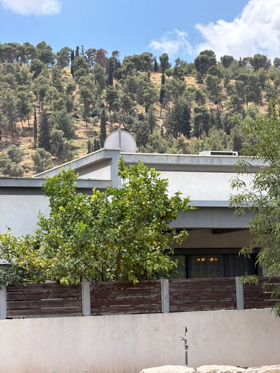 The image shows a building in the Gilboa Regional Council, Israel, with a hillside covered in trees in the background. The building has a modern design with a white exterior and wooden accents. A tree with green leaves is growing near the building. The sky is blue with some clouds.