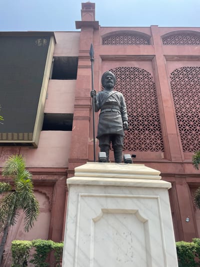 The image shows a statue of a man holding a spear, standing on a white marble pedestal. The statue is located in front of a large, red sandstone building with intricate lattice work on its upper facade. A large, dark electronic screen is visible on the left side of the building, partially obscuring it. Palm trees and green bushes are present in the foreground and to the left of the building. The sky is bright and clear. This is a view of the statue of Baba Deep Singh at the entrance to the Golden Temple complex in Amritsar, India.