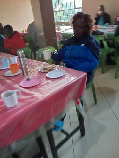 The image captures an indoor scene, likely a dining hall or cafeteria, where several individuals are gathered for a meal or refreshments. In the foreground, a woman with reddish-brown, textured hair, wearing a blue and black jacket, is seated at a table covered with a translucent pink plastic tablecloth. She holds a white mug in her right hand, suggesting she is drinking. Her face is slightly blurred. Her feet, clad in light blue open-toed shoes, are visible beneath the table.
On the table in front of her are two slices of bread on a white plate, alongside an empty white plate and a pink plate. Other items on the table include a silver thermos flask, and various mugs in white, light blue, and orange colors. In the background, other people are visible sitting at additional tables, also covered with similar pink plastic tablecloths, and using green plastic chairs. One person in a red hoodie is seen to the left. A large window with a metal grille allows natural light into the room, indicating it is daytime. The overall atmosphere appears casual and communal. No text is visible in the image. The scene is set in what is stated to be Riara Ridge, Kenya.