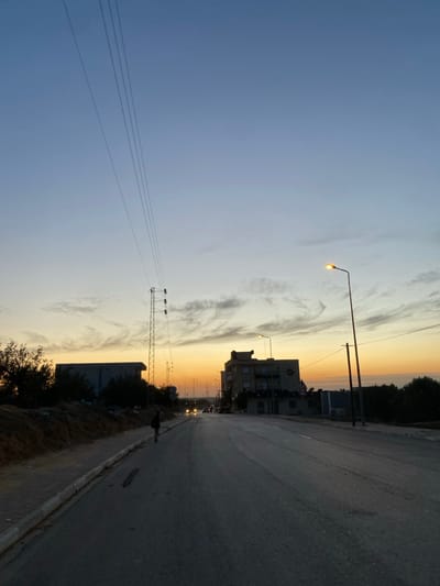 A street scene at dusk. The sky transitions from blue to yellow-orange near the horizon. A streetlamp glows in the fading light. A building and utility poles line the roadside. A paved road stretches into the distance. The overall image is a landscape dominated by the sky and the street scene elements.