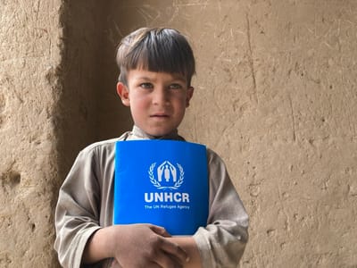 A young boy is standing in front of a mud brick wall. He is wearing a light brown shirt and holding a blue pamphlet with the UNHCR logo and the words "The UN Refugee Agency" printed on it. The boy's facial expression is neutral, and he is looking directly at the camera. The scene is located in Saranan, Pakistan (latitude: 30.532, longitude: 66.909). The background is a simple mud brick wall with a few scratches on it. The boy's hands are holding the pamphlet firmly. The photograph is taken from a slightly angled view, with the boy positioned in the center of the frame. The lighting is natural and slightly diffused.