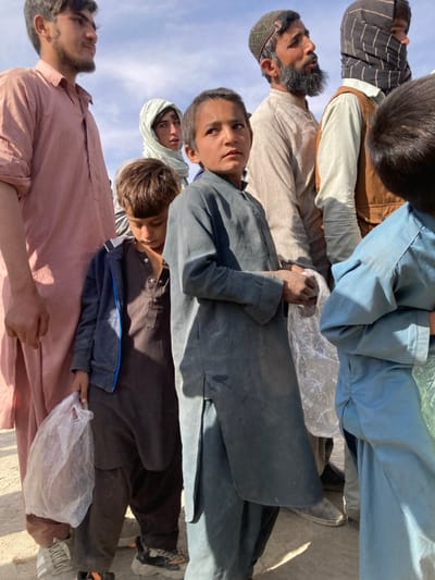 A group of approximately seven individuals, primarily males of varying ages and one partially visible woman, stand outdoors in what appears to be a queue or gathering in Chaman City Tehsil, Pakistan. The scene is illuminated by strong sunlight from the upper right, casting distinct shadows. The ground is a light-colored dirt or dust.
In the foreground, a young boy wearing a dark gray traditional tunic and trousers stands facing slightly right, looking upwards. His hands are clasped at waist level, appearing to hold a clear plastic bag. To his immediate left, another younger boy in a brown tunic, trousers, and an open blue hooded jacket looks downwards, holding a clear plastic bag in his left hand. To the far left, a young man with dark hair and light facial stubble wears a faded pinkish-brown traditional tunic and trousers, looking to the right. He wears white sneakers with black stripes.
Behind the central young boy and to his left, a woman is partially visible, wearing a light-colored head covering and looking towards the right. To the right of the central boy, an adult man with a dark beard and a patterned skullcap wears a light beige traditional tunic and trousers, looking upwards and to the right. His footwear consists of dark, traditional shoes. Behind this man, a second adult male is partially visible, wearing a brown vest over a light garment and a dark patterned head covering, facing right. To the far right, a boy in a light blue traditional tunic and trousers is visible from the back, holding a clear plastic bag. All visible individuals are dressed in traditional South Asian attire.