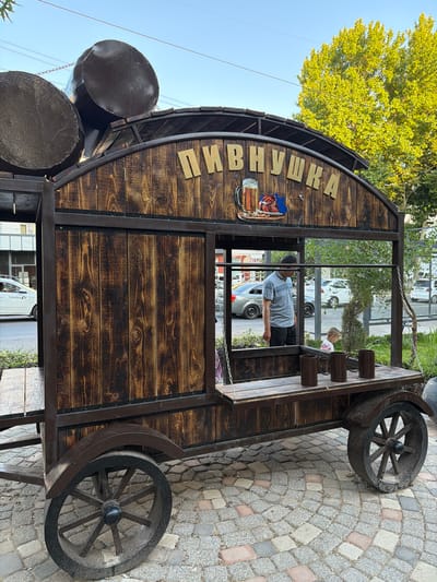 In Chirchik, Uzbekistan, a wooden cart with a rustic design resembling barrels is parked on a cobblestone pavement. The cart is adorned with the Cyrillic inscription "ПИВНУШКА" in gold lettering, accompanied by a graphic of a beer mug and food. Inside the cart, a man in a striped shirt and dark pants is visible, as well as a young child. The cart has large wooden wheels and is situated outdoors, with a street and buildings in the background. The scene is illuminated by natural daylight, suggesting it is daytime.