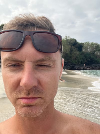 The image is a selfie taken by a man on a beach in Manggis, Indonesia. The man is wearing sunglasses and is looking at the camera. The background shows a beach with waves and lush green vegetation on a rocky cliff. The sky is overcast, suggesting it might be late afternoon or early morning. The overall mood appears relaxed and casual. The man's face is close to the camera, partially obscuring the background. The water is clear and the sand is light-colored. There are a few other people visible in the distance on the beach and in the water, enjoying the scenery. The image is taken with a vertical orientation.