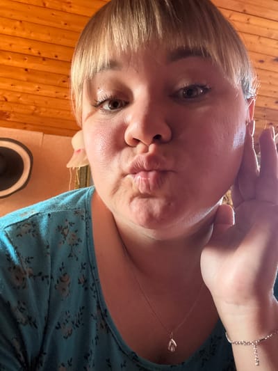 A woman with blonde hair and bangs is taking a selfie. She is wearing a blue shirt with a floral pattern and a silver necklace with a teardrop pendant. Her hand is resting on her cheek and her lips are pursed. The background is a wooden wall. The image appears to have been taken indoors, possibly in a home in Nizhne-Chufichevo, Russia.