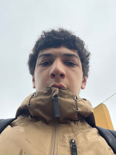 The image presents a low-angle, close-up portrait of a young adult male facing directly towards the camera. He possesses dark, curly hair and dark eyes. His mouth is slightly open, with his lower lip partially obscured by his clothing. He is attired in a light brown or tan jacket, zipped up with its collar elevated to cover his chin. The jacket features a central zipper with a black pull tab, metallic eyelets, and dark fabric elements on the collar's drawstring. Apparent dark smudges or stains are visible on the upper left portion of the jacket. A black strap, consistent with a backpack, is visible over his right shoulder. The background is dominated by an overcast, light gray sky, with a narrow section of a yellow or light brown architectural structure visible on the far right. This outdoor scene is stated to be located in Meistratzheim, France.