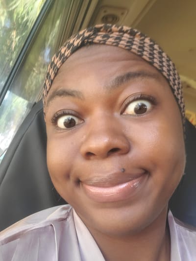 A close-up, selfie-style photograph shows a Black woman with a wide, surprised expression, her eyes looking directly at the camera. She is wearing a headscarf with a black and tan houndstooth pattern and a light-colored, silky top. A small mole is visible on her upper lip, just above the center. The background shows a car interior, with a dark seat on the right and a window with greenery on the left, suggesting the photo was taken while traveling. The lighting appears to be natural daylight. The image is consistent with a personal photograph taken in Gwagwada, Nigeria, a location known for its residential and semi-urban characteristics.
