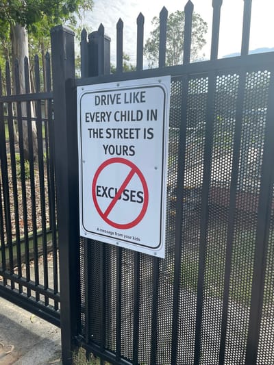 A white rectangular sign with a safety message for drivers is prominently displayed on a black metal fence in Riverhills, Australia. The sign, which is oriented vertically, features bold black text on a white background. The main message reads: "DRIVE LIKE EVERY CHILD IN THE STREET IS YOURS." Below this, a large red circle with a diagonal line through it, similar to a "no" symbol, encompasses the word "EXCUSES," conveying "NO EXCUSES." In smaller text at the bottom right, it states, "A message from your kids."

The sign is attached to a sturdy black metal fence, which appears to have pointed finials along its top edge and a section of perforated mesh panels, visible in the upper part of the image. The lighting suggests it is daytime with a clear or partly cloudy sky. In the background, beyond the fence, various trees and green foliage are visible, indicating an outdoor, possibly suburban or park-like setting. The overall scene suggests a residential area where child safety is a concern.