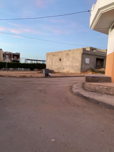 The scene depicts an exterior urban environment in الغنادة, Tunisia, during twilight hours. A dark, granular paved road occupies the foreground, curving slightly towards the right. A concrete curb and an elevated sidewalk segment are visible along the right edge of the frame. In the mid-ground, a broad, unpaved dirt lot contains a grey, rectangular utility container. To the left, a multi-story residential building with light-colored walls and dark accents is partially obscured by a dense, uniform green hedge. Behind the hedge line, a long, low-profile structure with a flat roof is discernible. To the right of the dirt lot stands a single-story, grey concrete building, exhibiting characteristics of ongoing construction or an unfinished state, featuring a small square window opening and a larger rectangular void. The upper right corner reveals a partial view of a light-colored building with an orange-painted section. The sky overhead is light blue with faint pink and orange cloud formations, traversed horizontally by multiple black utility lines. No individuals, actions, or specific interactions are observed within the frame.