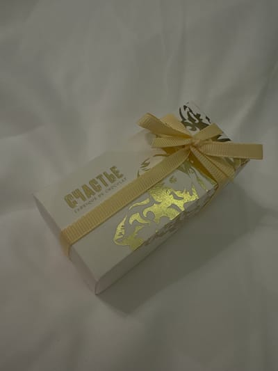 A small, white, rectangular gift box with golden filigree patterns is centrally placed on a slightly off-white, possibly fabric surface. The box is tied with a light yellow ribbon bow. The label on the side of the box reads "Счастье Fabrique de Chocolat" in gold lettering. There is no indication of the city of Sertyakino or any people in the image.
