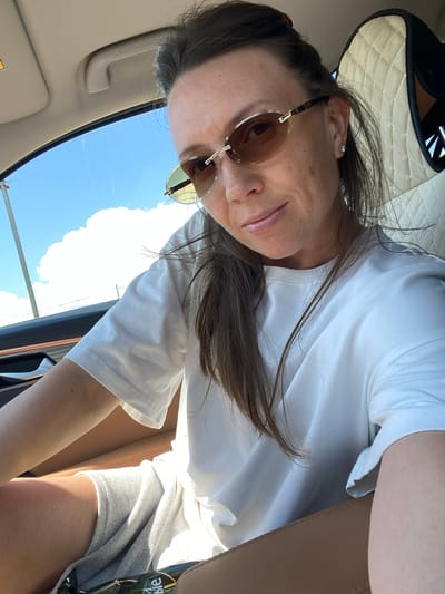 A woman is sitting in the driver's seat of a car. She is wearing sunglasses and a white t-shirt. The car's interior is visible, with a tan leather seat and quilted headrest. Outside the car, the sky is blue with a large white cloud. The location is identified as Kvards, Russia, which is a village in the Kurchaloyevsky District of the Chechen Republic, Russia.