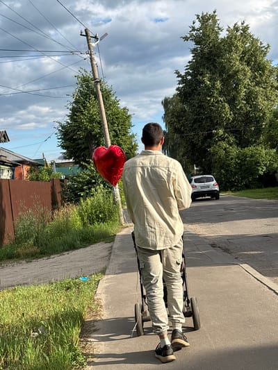 A person is walking away from the camera down a paved path beside a road, pushing a stroller. The person is wearing a light-colored, long-sleeved shirt and light-colored cargo pants, with dark sneakers and white socks. They are holding a red, heart-shaped balloon on a string. To the left of the path is a wooden fence and some vegetation, with houses visible beyond the fence. To the right of the road, there are trees and a white car is driving away from the camera. Overhead, power lines crisscross the sky, which is partly cloudy. The scene appears to be in a residential area.