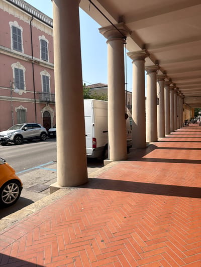 The image depicts a street scene in Santarcangelo di Romagna, Italy, characterized by a long colonnade with prominent rounded columns. The colonnade casts distinct shadows on the red brick pavement, laid in a herringbone pattern. In the foreground, the wheel and lower portion of a small orange car are visible on the left. Behind it, a silver SUV is parked on the street, alongside a white van. The buildings lining the street are visible beyond the colonnade, featuring a pink facade with shuttered windows and decorative architectural elements. The sky is clear and blue, indicating a sunny day. In the distance, at the end of the colonnade, several parked motorcycles and bicycles suggest a public area.