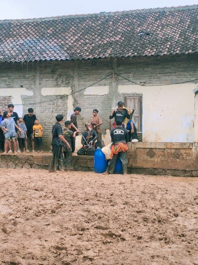A group of people, mostly men, are standing in front of a brick building in Kecamatan Kedawung, Indonesia, located at coordinates -7.494, 111.07. Some are talking while others are standing around or looking at something off-camera. The building has a tiled roof and there is mud on the ground in front of it. Some of the people are wearing black shirts with yellow writing. A blue barrel is in front of the building. The building is situated on a road, and there is a muddy area in front of it.  A few trees are visible in the background. The sky is gray and overcast. There is a possible rain event that took place recently. The people in the foreground are interacting with each other. It is likely they are waiting for something or about to participate in an event.  The scene is taken from a street level perspective. The people are standing on a paved surface. The weather is overcast and there are some small puddles of water on the ground.